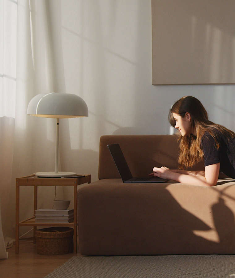 Kobieta pracująca na laptopie leżąc na brązowej sofie. Obok sofy stolik kawowy z książkami oraz dużą lampą z kloszem w kształcie grzyba.