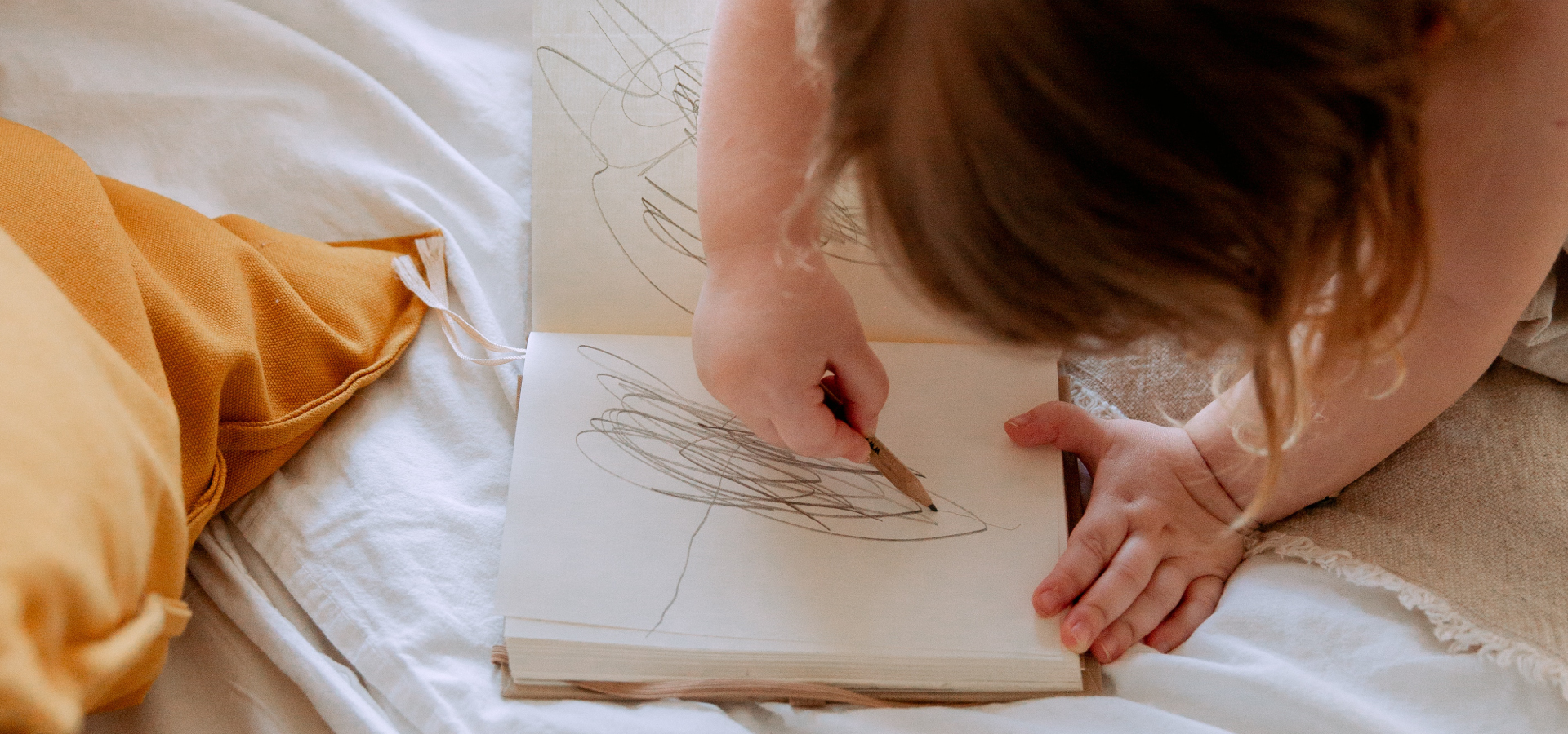 Zbliżenie na ręce dziecka, które rysuje w notatniku leżącym na łóżko. W tle żółta poduszka.
