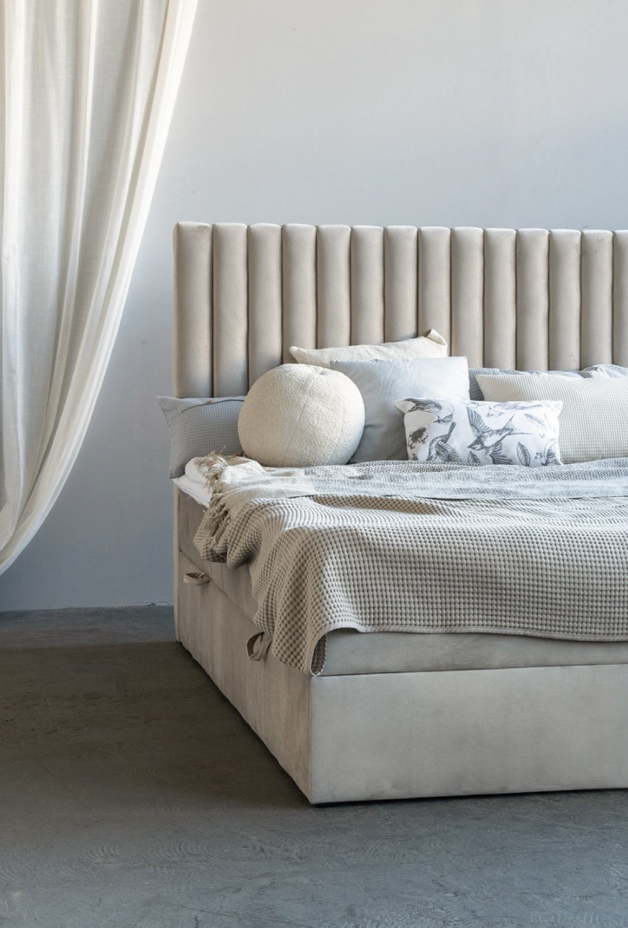 Welurowe, kontynentalne łóżko w jasnobeżowej tkaninie, na którym leży jasno-szara pościel oraz okrągła poduszka w kształcie piłki.
