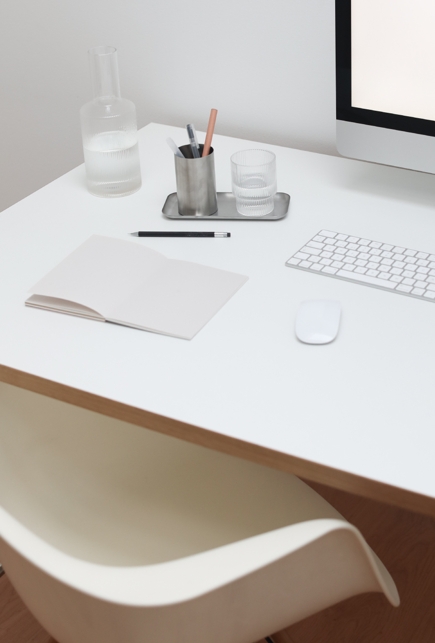 Białe biurko w stylu minimalistycznym z białym fotelem z tworzywa sztucznego. Na biurku notatnik oraz akcesoria komputerowe.