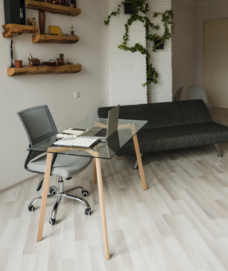 Wnętrze pomieszczenia z rozkładaną, grafitową kanapą, szklanym biurkiem na drewnianych nóżkach oraz obrotowym krzesłem w szarym odcieniu.