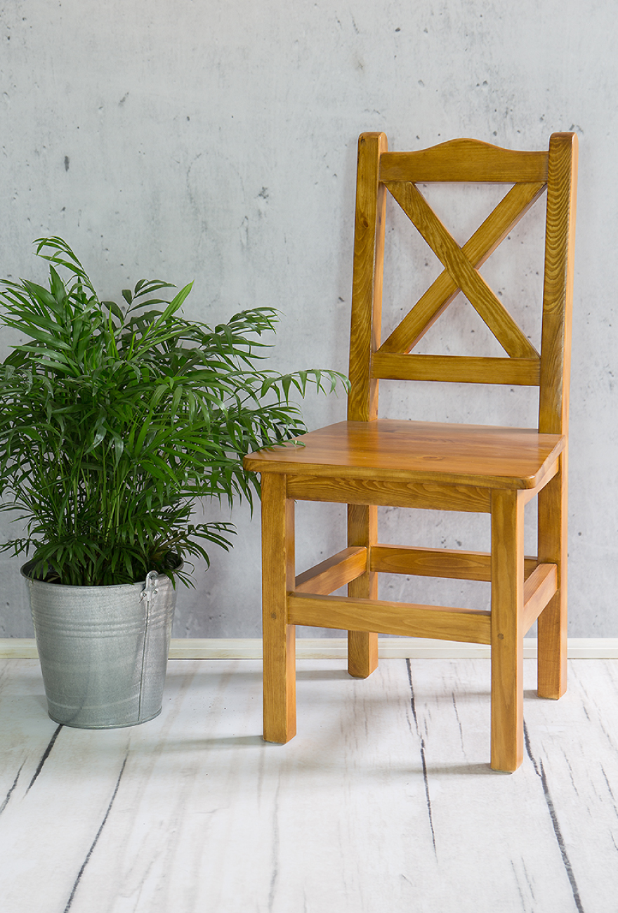 Drewniane krzesło w rustykalnym stylu stojące na tle szarej ściany, po lewej stronie zielona roślina w metalowej donicy.