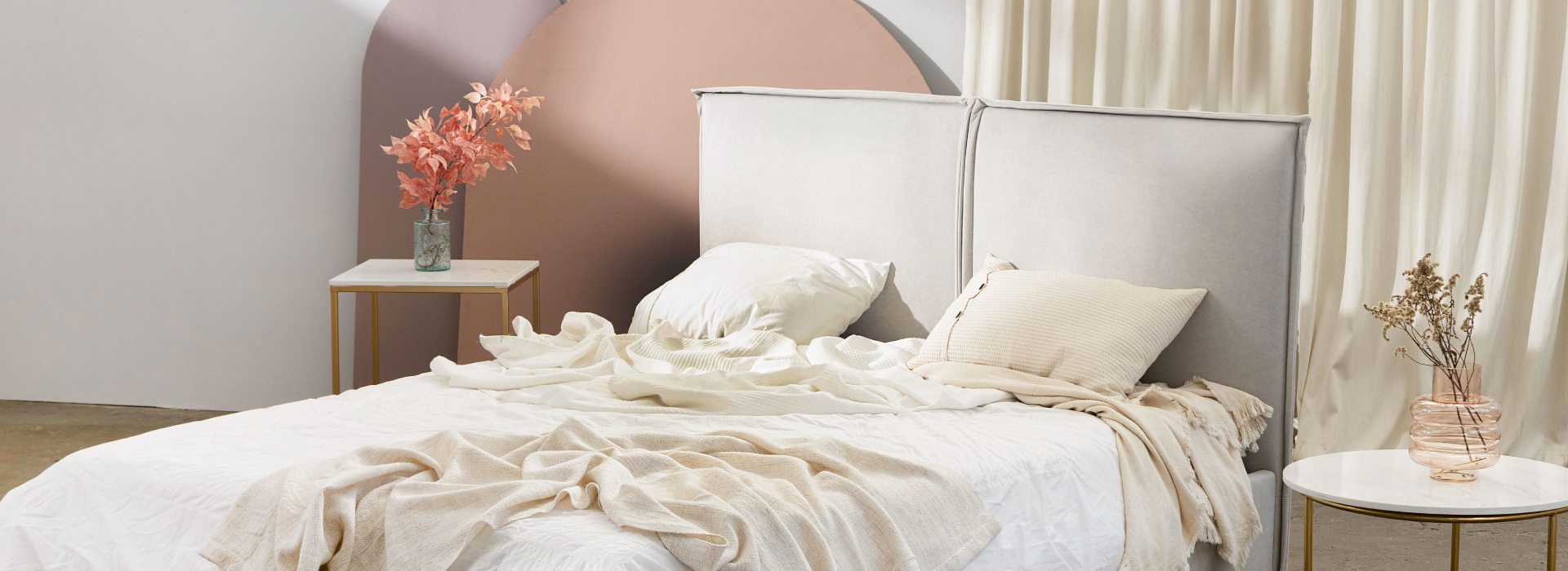 Welurowe łóżko o prostej formie w kolorze jasnoszarym. Na łóżku beżowo-biała pościel, po obu stronach łóżka stoliki kawowe.