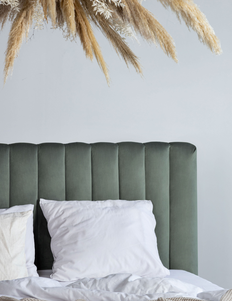 Welurowe łóżko z wysokim zagłówkiem w pionowe przeszycia, całość w kolorze pistacjowym. Na łóżku biała pościel i beżowa narzuta.
