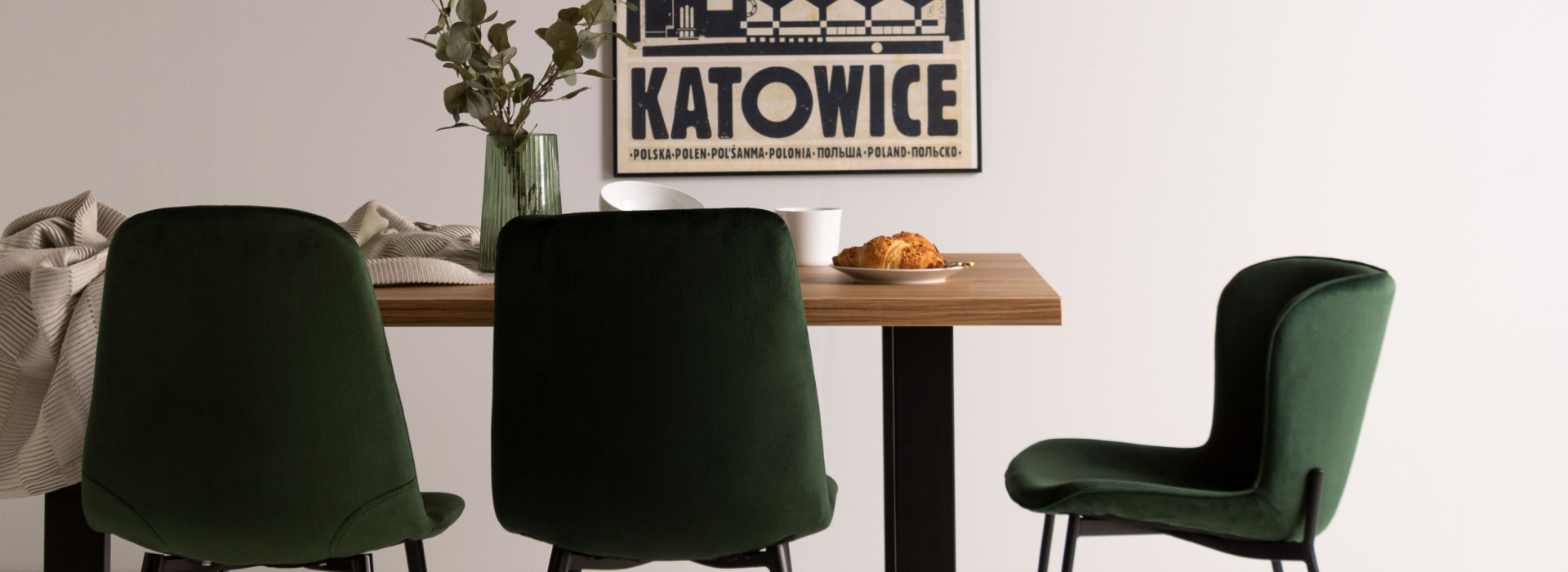 Butelkowo-zielone krzesła na czarnych nogach stojące wokół drewnianego stołu na czarnej konstrukcji. Na stole zastawa stołowa oraz zielony szklany wazon, w tle plakat z napisem „Katowice”.