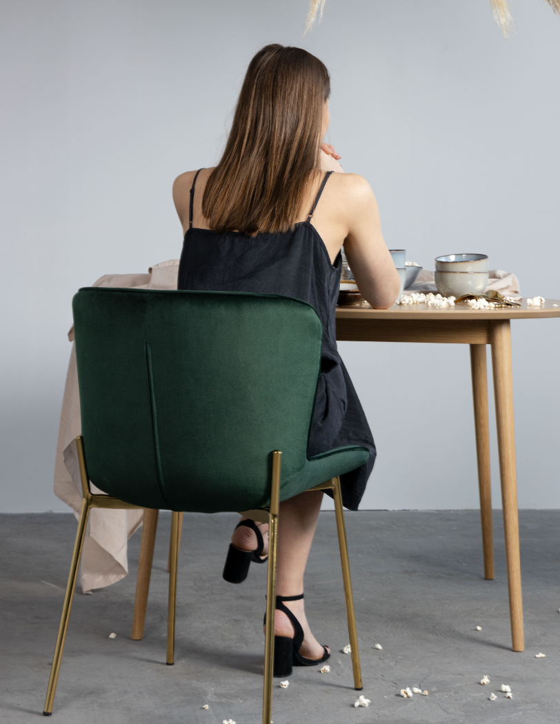 Butelkowo zielone krzesło na złotej podstawie, które stoi przy drewnianym stole. Na krześle siedząca tyłem dziewczyna, na stole zastawa.