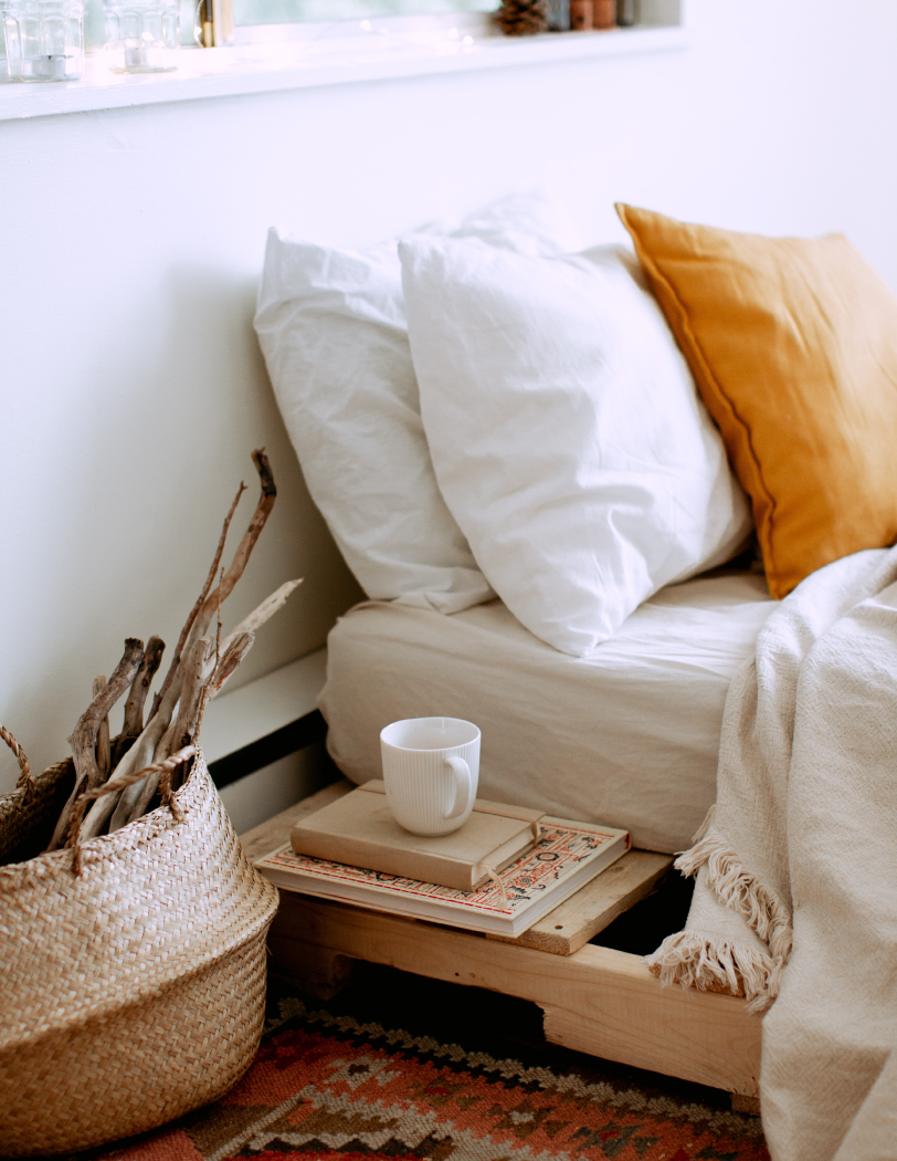 Wnętrze sypialni ze zbliżeniem na łóżko, na którym widoczna jest biało-pomarańczowa pościel. Przy łóżku niski stolik nocny, na którym leżą książki oraz kubek.