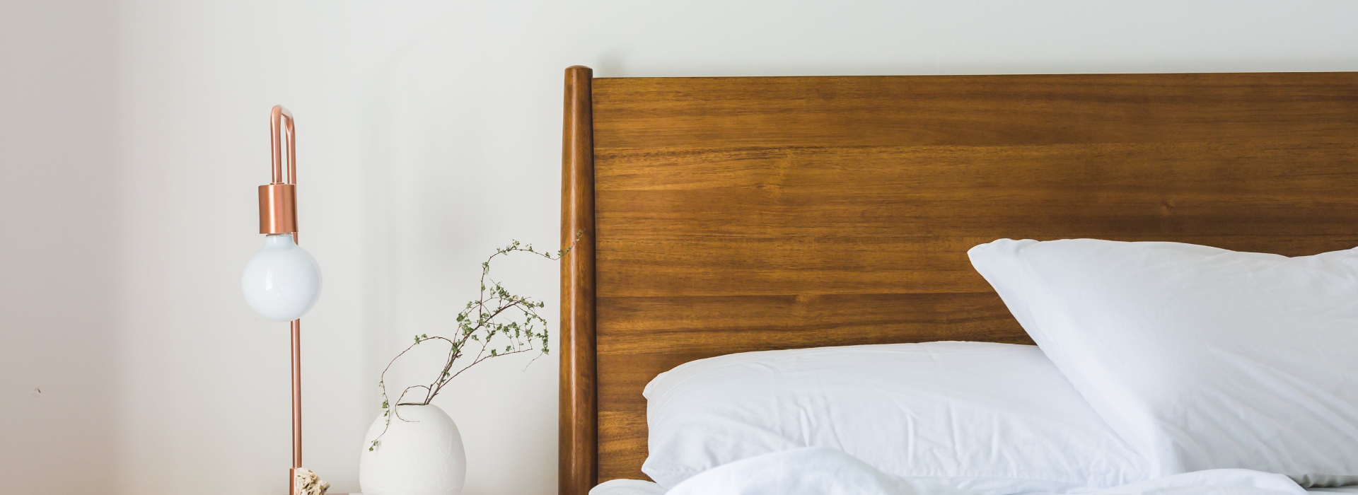Zbliżenie na drewniane łóżko, na którym leży biała pościel. Po lewej stronie widoczna szafka nocna z marmurowym blatem, na której stoi lampka oraz mały wazon z gałązkami.