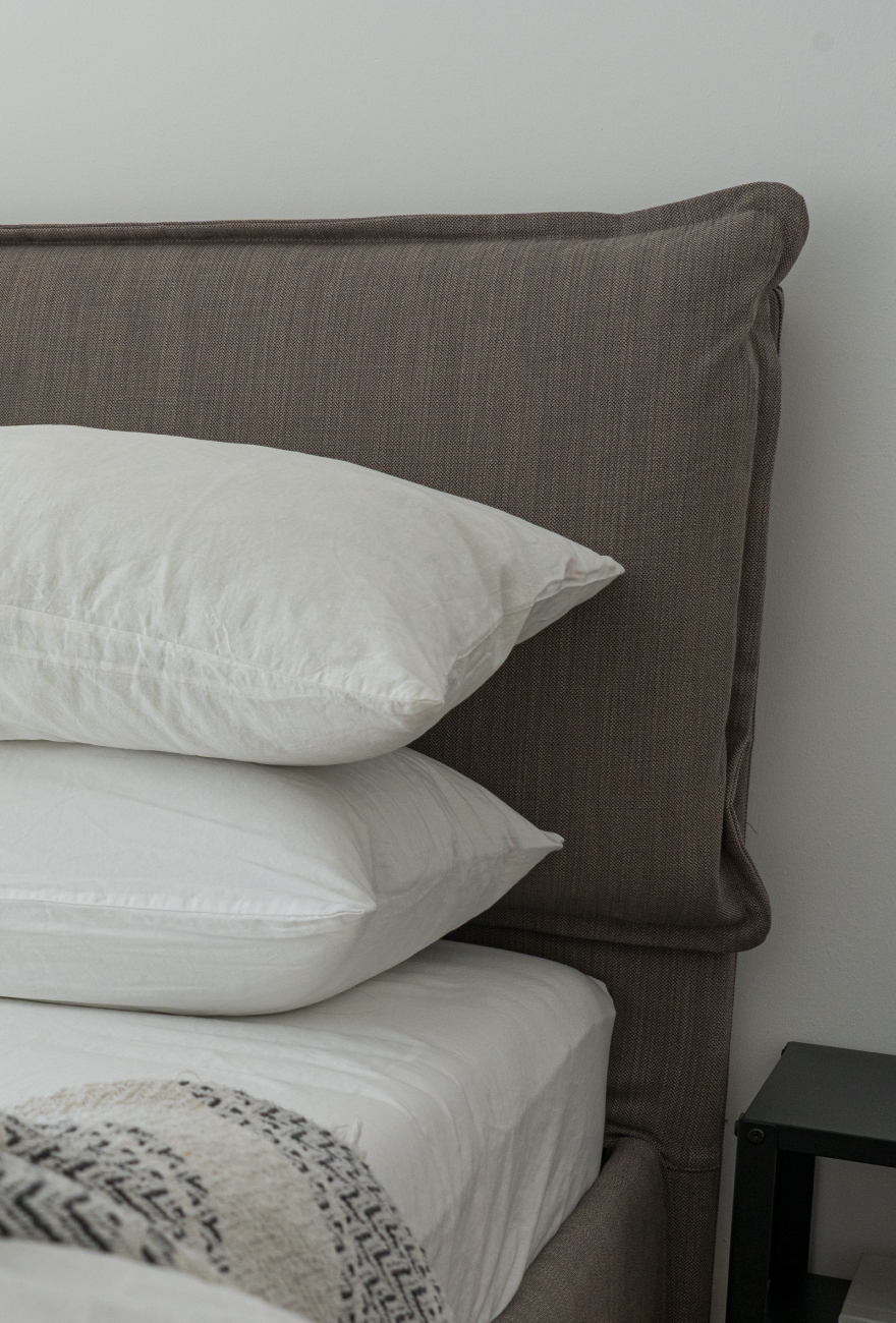 Zbliżenie na szare tapicerowane łóżko, na którym leżą poduszki i pościel. Po prawej stronie fragment czarnego stolika kawowego.