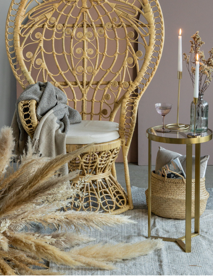 Wysoki rattanowy fotel w stylu boho z zaokrąglonym i zdobnym zagłówkiem. Po prawej stronie wysoki złoty stolik, na którym stoją świece oraz kieliszek.