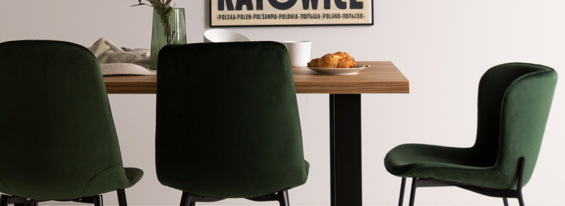 Butelkowo-zielone krzesła ustawione wokół drewnianego stołu, na którym stoi zastawa. W tle, na ścianie, zawieszony plakat z napisem „Katowice”.