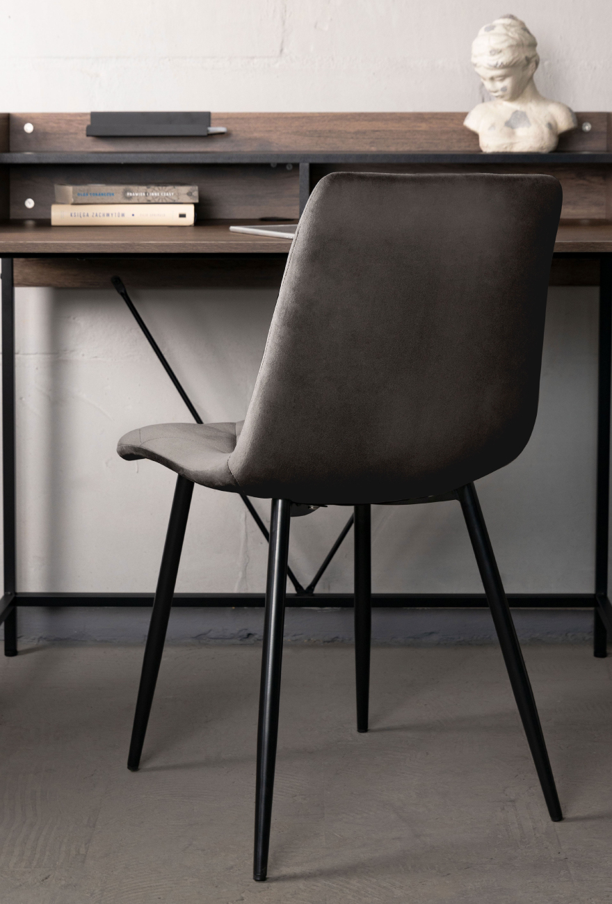 Szare welurowe krzesło na czarnych nogach stojące przed biurkiem w stylu industrialnym.
