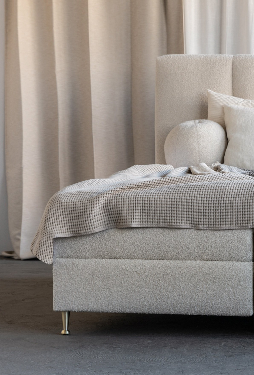 Łóżko kontynentalne w tkaninie boucle z pionowymi przeszyciami na zagłówku. Na łóżku ułożone poduszki oraz narzuta.