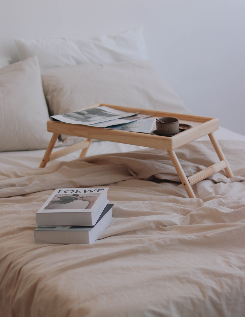 Łóżko z beżową pościelą, na której stoi drewniana taca. Na tacy leżą książki oraz filiżanka z kawą. Obok tacy, na pościeli, leżą dwie kolejne książki.