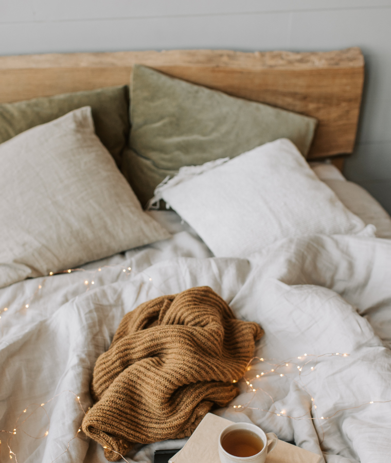Łóżko z dekoracyjnie ułożoną pościelą i poduszkami, na których leży książka, kubek herbaty oraz karmelowy koc.