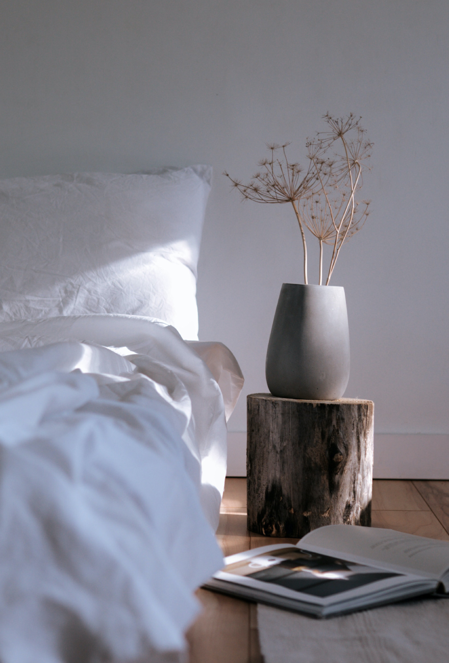 Niskie łóżko z białą pościelą, obok którego stoi stolik nocny wykonany z pnia drzewa. Na nim stoi szary wazon z suszkami.