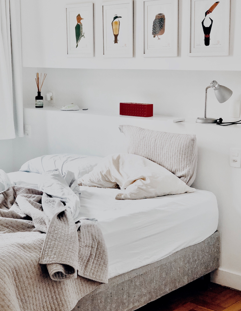 Szare łóżko z białą pościelą, na tle białej ściany z kolorowymi obrazkami papug i innych ptaków.