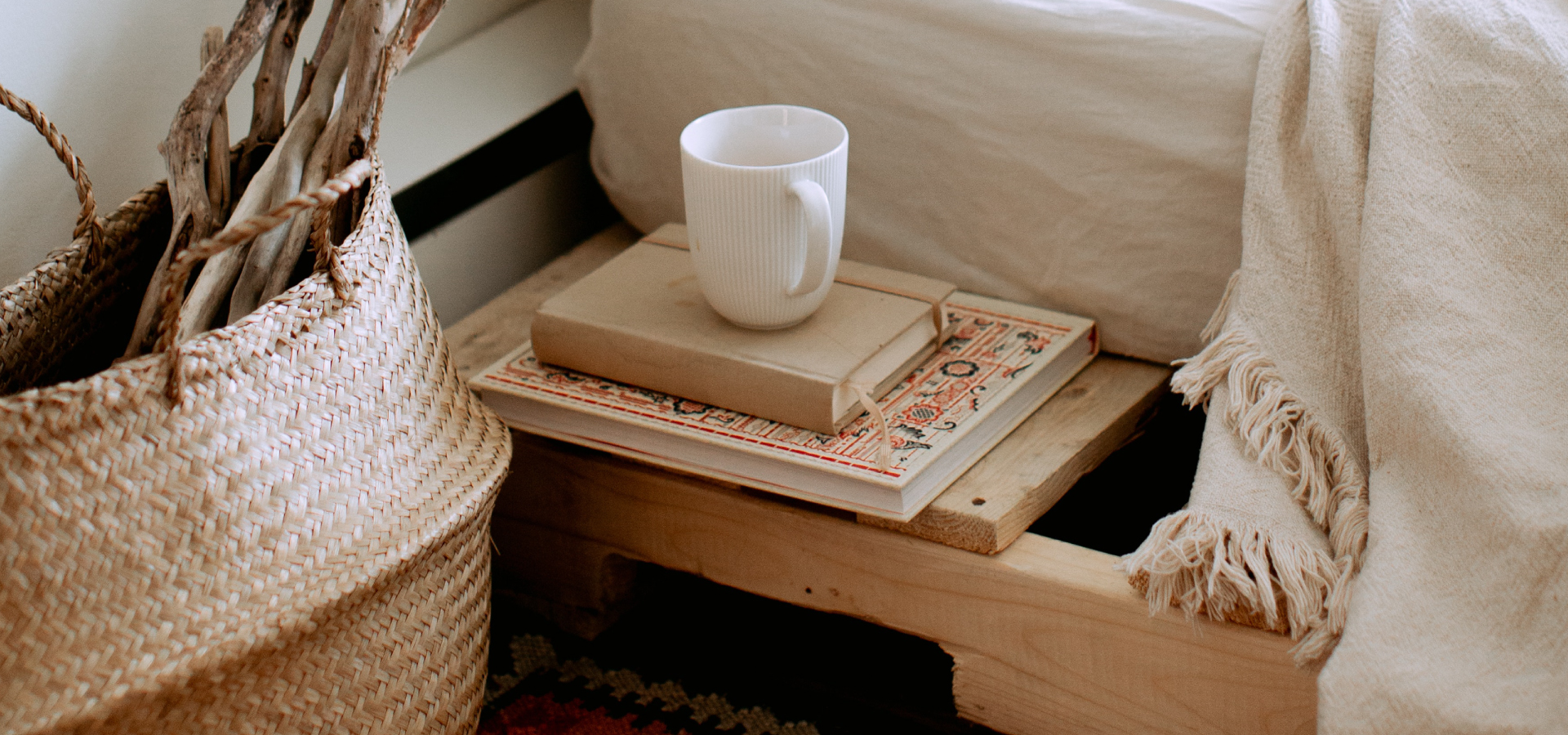 Zbliżenie na bok łóżka z białą pościelą, obok łóżka niski stolik nocny z książkami i kubkiem. Po lewej stronie kosz wiklinowy z suszkami.