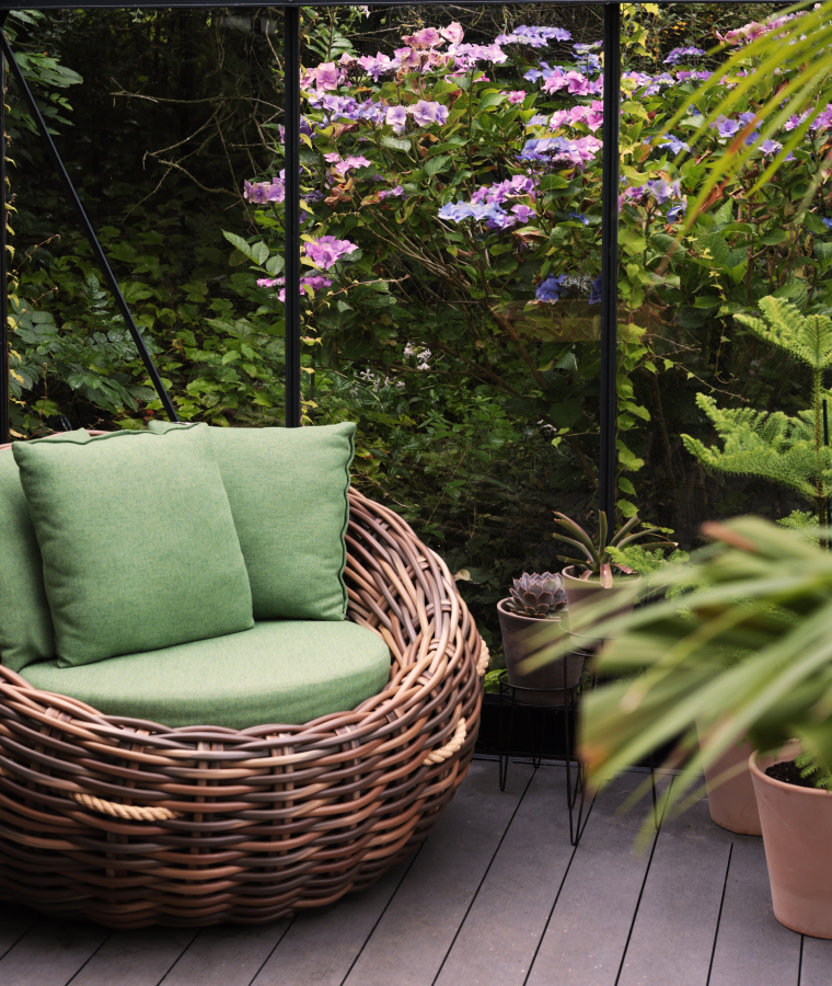 Wnętrze szklarni ogrodowej z duży, wiklinowym fotelem z zielonymi poduszkami, wokół którego stoją rośliny liściaste. Za szklarnią bujna roślinność ogrodowa.