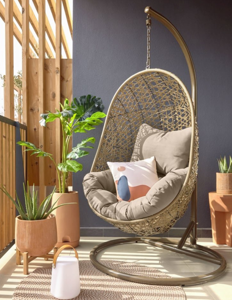 Fotel kokon o złoto-beżowym kolorze, który stoi na balkonie, wokół niego doniczki ceramiczne z roślinami.