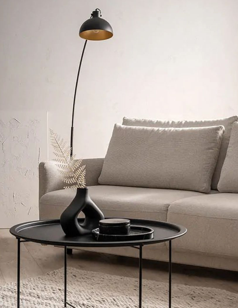 Beżowa sofa z dużym kompletem poduszek, przed którą stoi nowoczesny stolik w kolorze czarnym i z czarnymi dekoracjami. W tle wysoka lampa na długiej nodze oraz biały abstrakcyjny obraz.