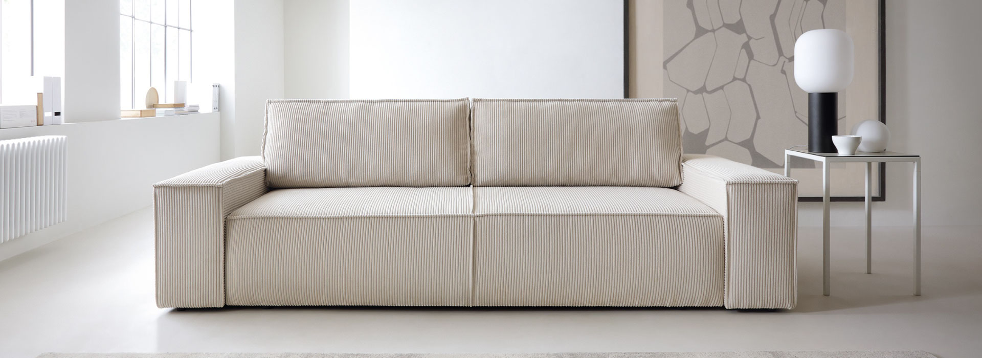 Jasnobeżowa sofa rozkładana o prostej i minimalistycznej formie w całości wykonana z tkaniny sztruksowej. Po prawej stronie stolik pomocniczy, na którym stoją dekoracje i lampa na czarnej nodze.