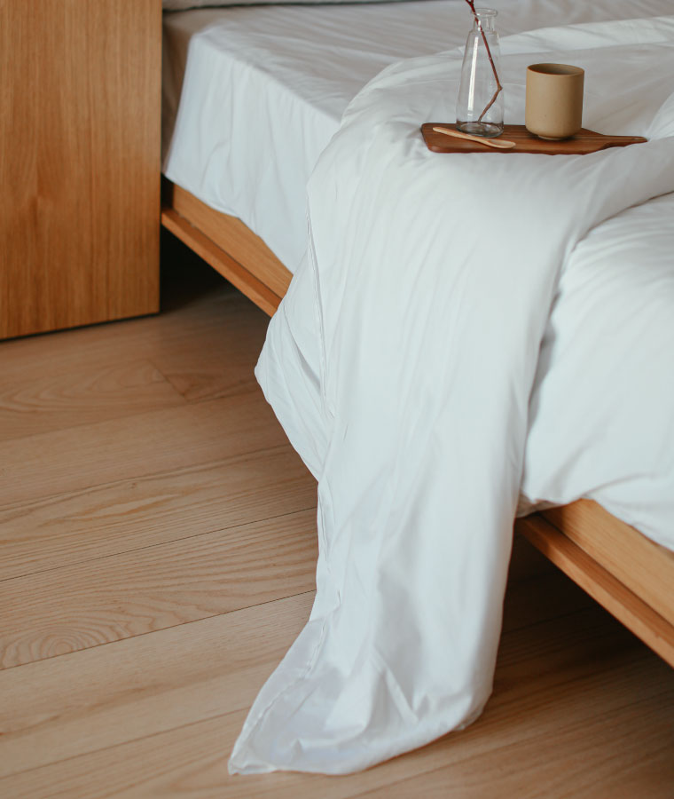 Zbliżenie na bok drewnianego łóżka, na którym leży biała pościel. Na pościeli mała drewniana tacka z kubkiem oraz szklanym wazonikiem.