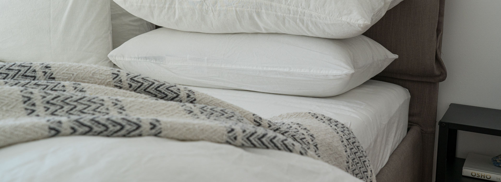 Zbliżenie na łóżko w kolorze szarym, na którym leżą dwie poduszki ułożone jedna na drugiej oraz beżowo-granatowy koc.
