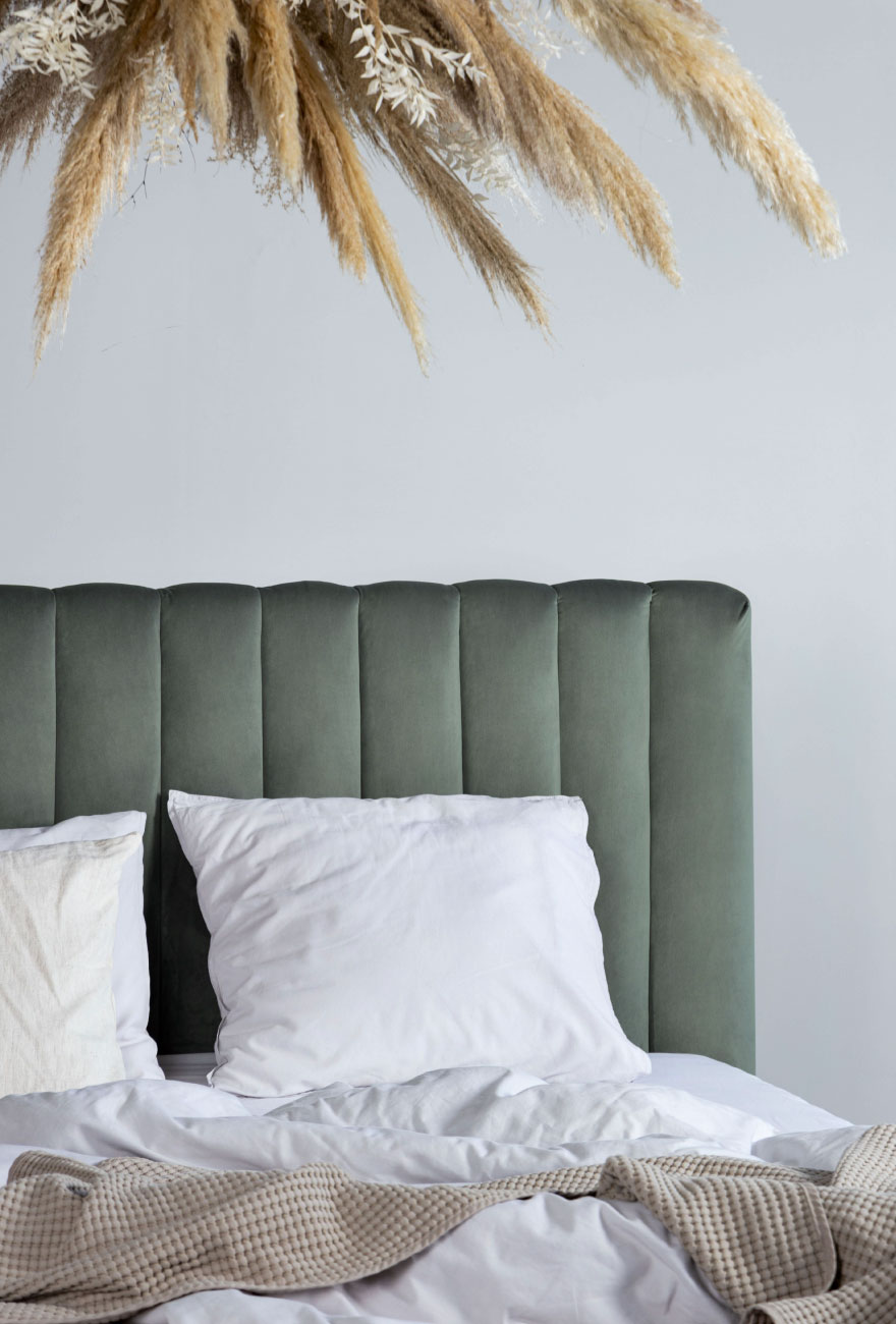 Zbliżenie na wysoki zagłówek łóżka w kolorze pistacjowym. Zagłówek z dekoracyjnymi przeszyciami w pionie, na łóżku biała pościel oraz beżowa narzuta.