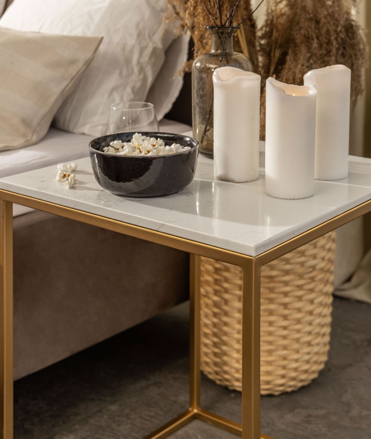 Wysoki stolik kawowy na złotej konstrukcji i marmurowym blacie, na którym stoją świece oraz miseczka z popcornem, w tle beżowe łóżko.
