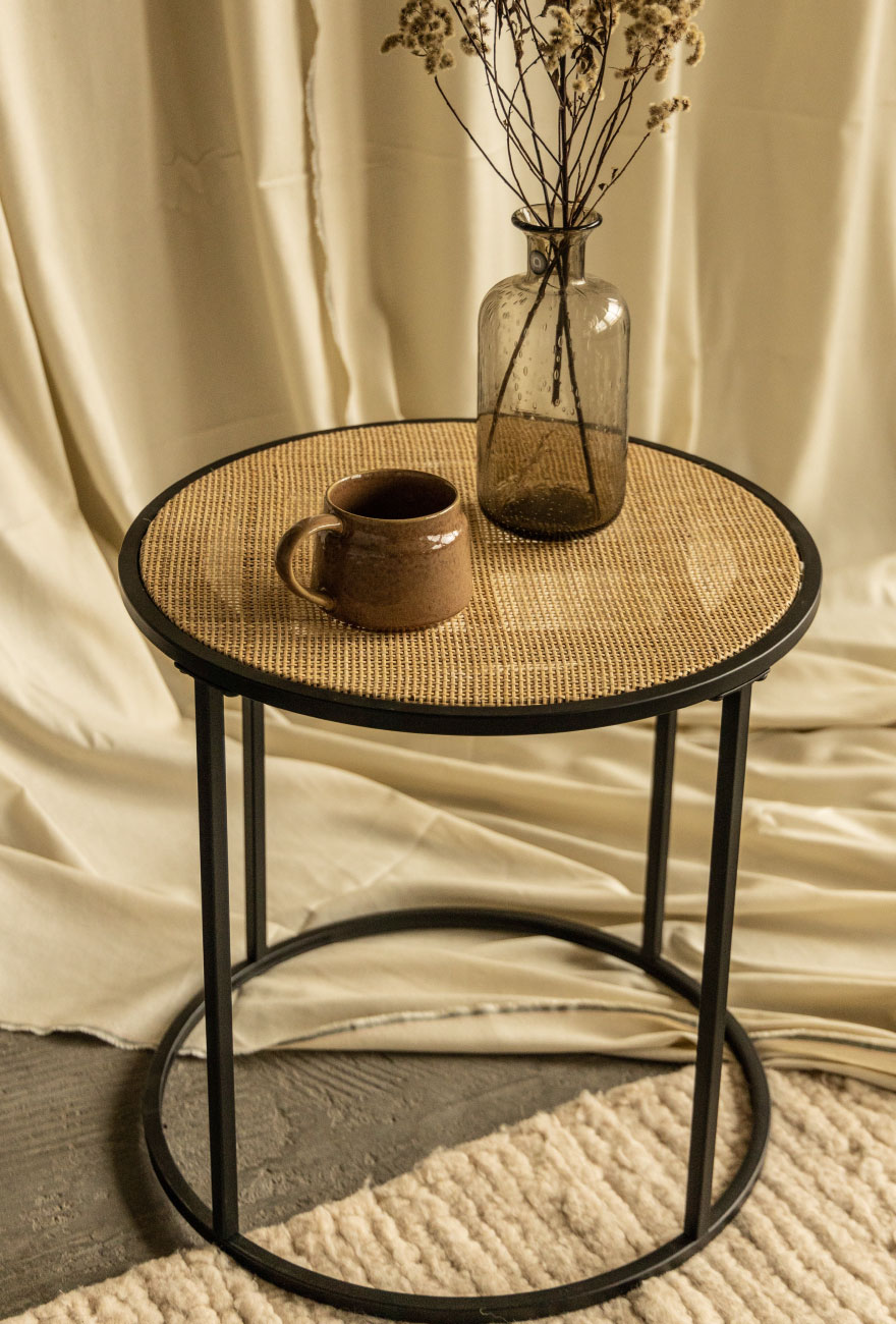 Stolik kawowy na metalowej okrągłej konstrukcji z rattanowym blatem, na którym stoi kubek oraz szklany wazon.