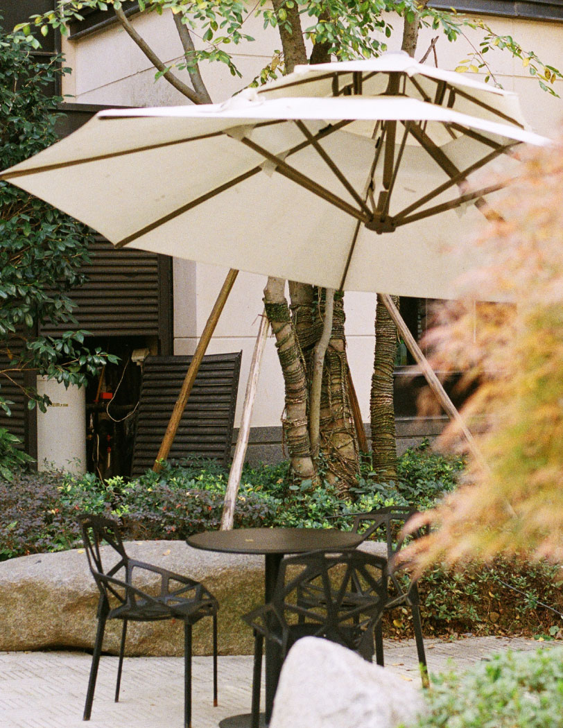 Ogródek wewnątrz dziedzińca z czarnym stolikiem oraz krzesłami, a także wysokim beżowym parasolem na drewnianej konstrukcji.