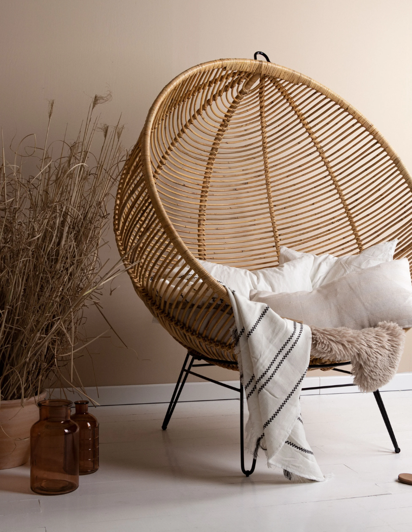 Okrągły fotel boho wykonany z naturalnego rattanu. Na fotelu ułożone poduszki i koc. Po lewej stronie fotela donica z trawami oraz dwa szklane wazony. Przed fotelem położone skórzane klapki w brązowym kolorze.