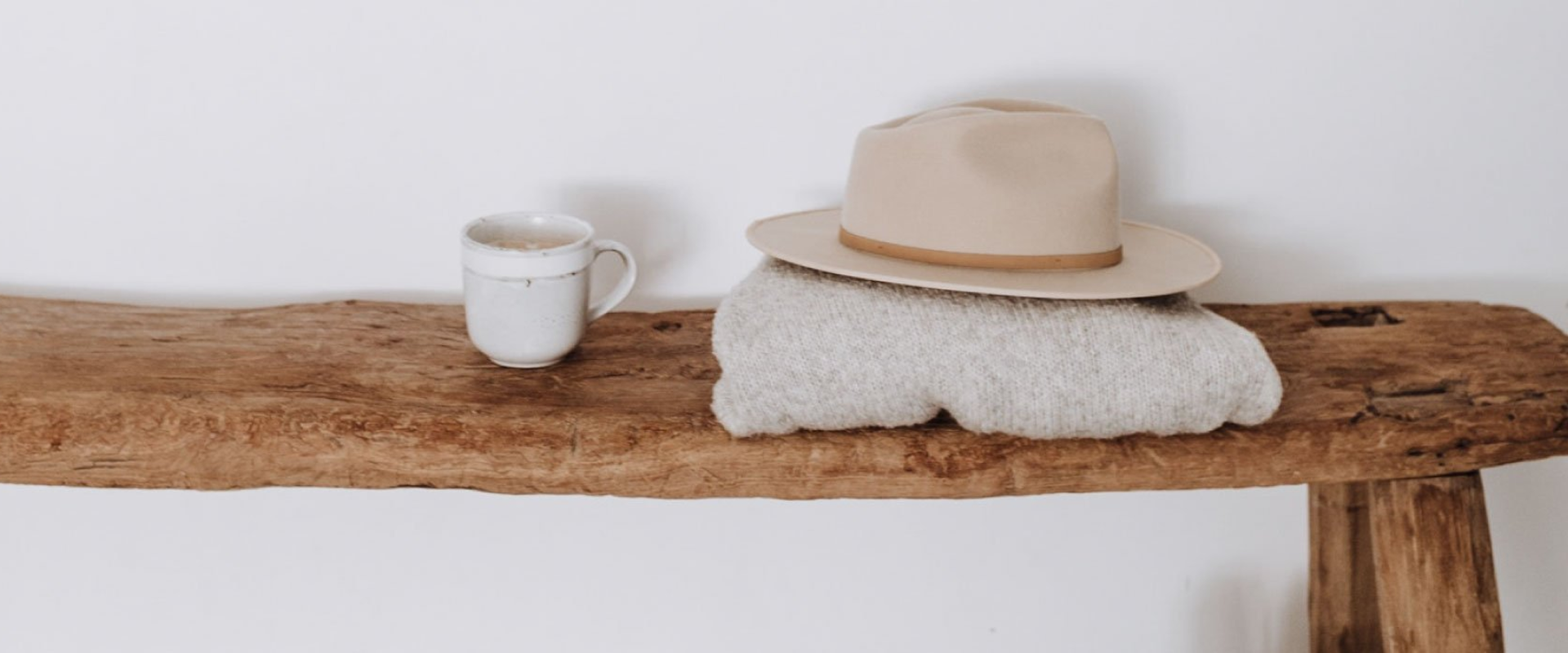 Rustykalna ławka z naturalnego drewna, na której stoi ceramiczny kubek w stylu vintage oraz leży złożony sweter z położonym na nim beżowym kapeluszem z rondem.