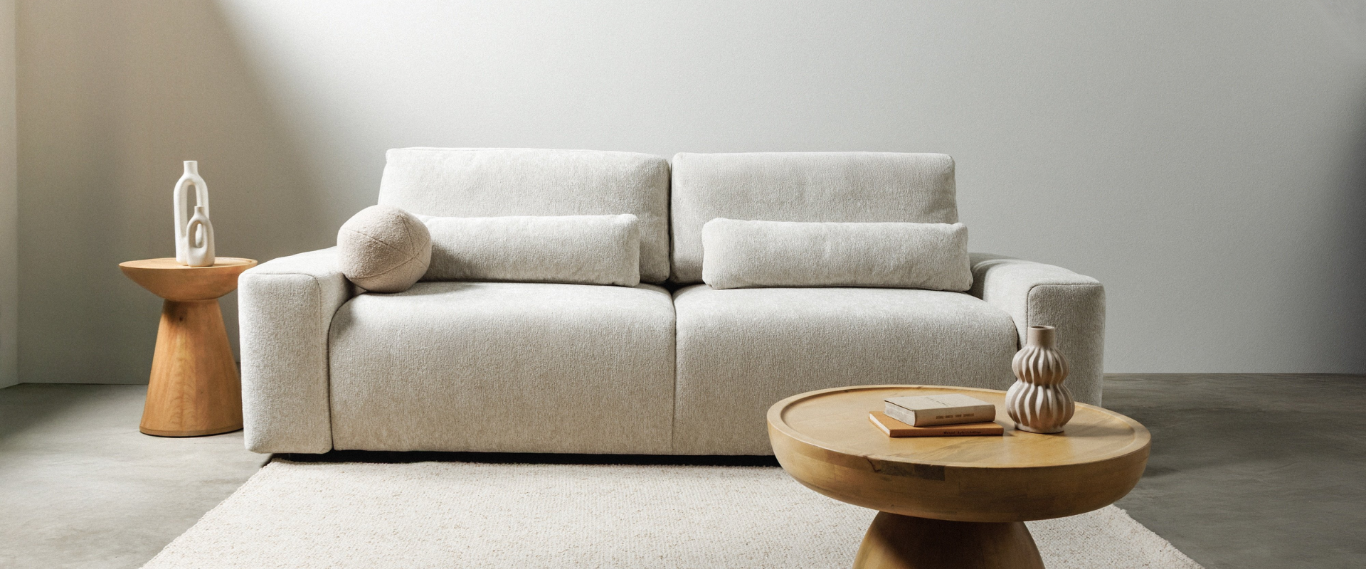 Minimalistyczny salon z beżową sofą w tkaninie boucle, ozdobioną podłużnymi i okrągłymi poduszkami, dwoma drewnianymi stolikami pomocniczymi z dekoracyjnymi wazonami oraz jasnym dywanem na betonowej podłodze.