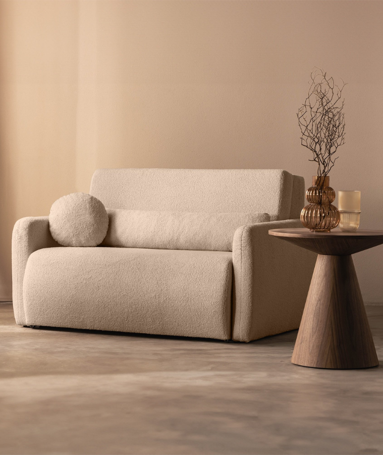 Beżowa sofa z tkaniny boucle w minimalistycznym wnętrzu. Obok znajduje się drewniany stolik w kształcie stożka z brązowym wazonem i suchą gałązką, tworząc przytulny, nowoczesny klimat.
