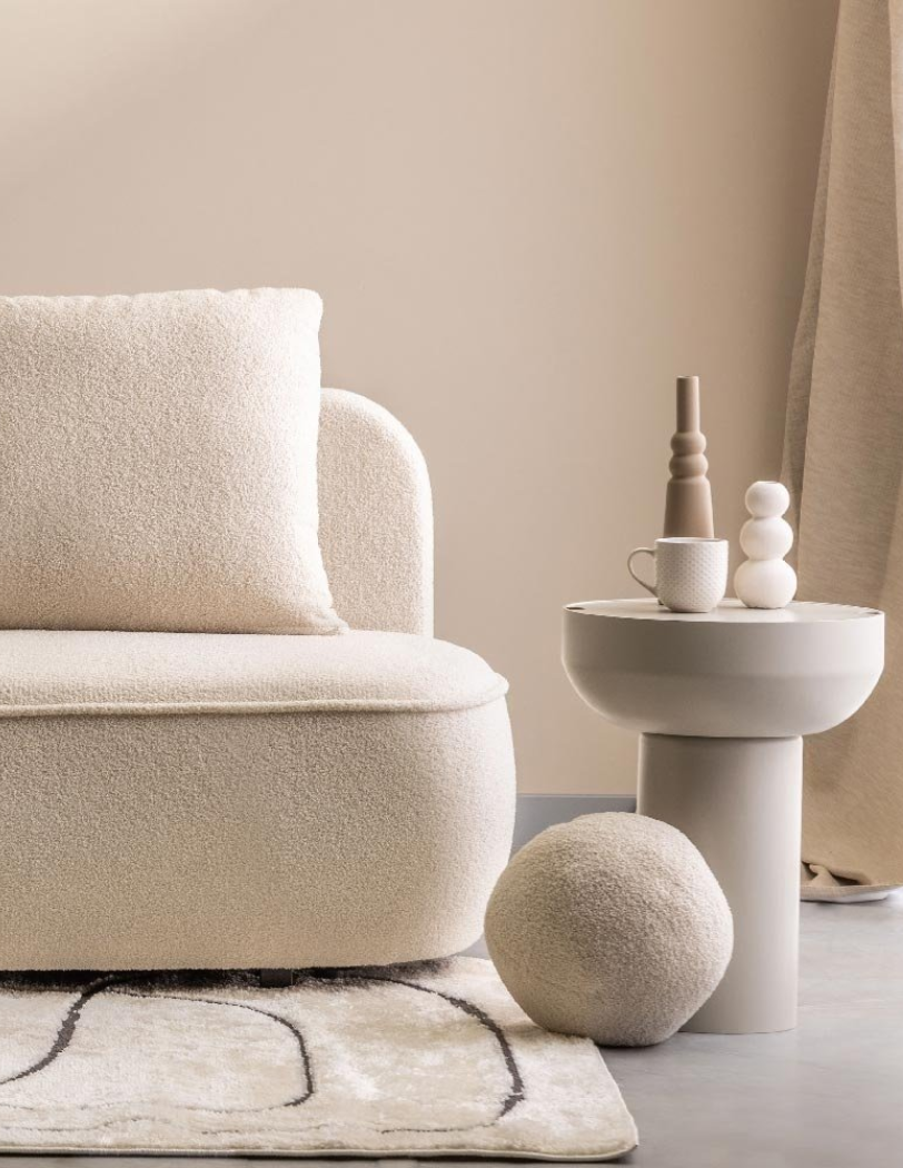Minimalistyczne wnętrze w odcieniach beżu – fragment sofy z miękkiej tkaniny boucle, obok okrągły stolik pomocniczy z ceramiką i filiżanką, a na podłodze dywan z geometrycznym wzorem i ozdobna kula.