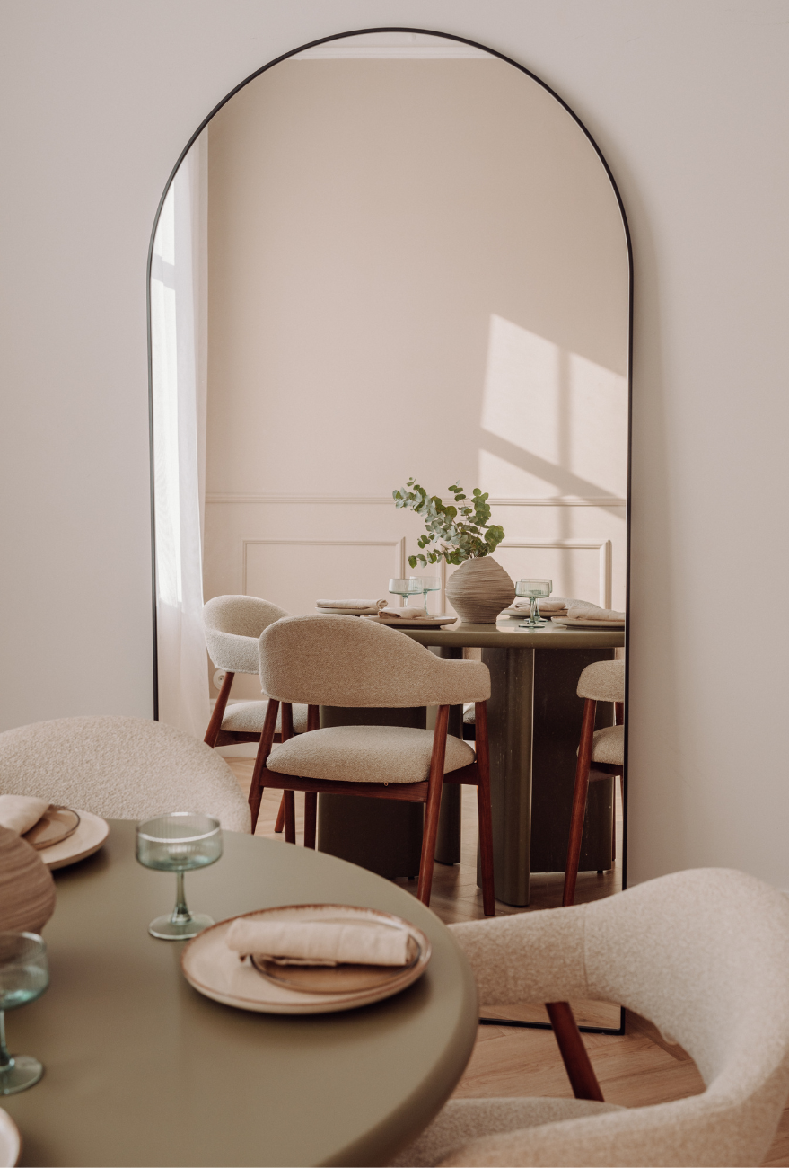 Elegancka jadalnia z okrągłym stołem w oliwkowym odcieniu i beżowymi krzesłami o drewnianych nogach, odbijająca się w dużym lustrze z czarną ramą. Na stole znajduje się wazon z zielonymi gałązkami i nakrycia w stonowanych barwach, tworzące harmonijną, przytulną atmosferę.