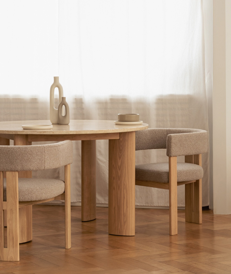 Jasne, minimalistyczne wnętrze z okrągłym stołem z jasnego drewna i dwoma tapicerowanymi krzesłami w beżowym odcieniu. Na stole stoją dwie dekoracyjne ceramiczne wazony i talerze, a w tle widoczne są lekkie, półprzezroczyste zasłony przepuszczające naturalne światło.
