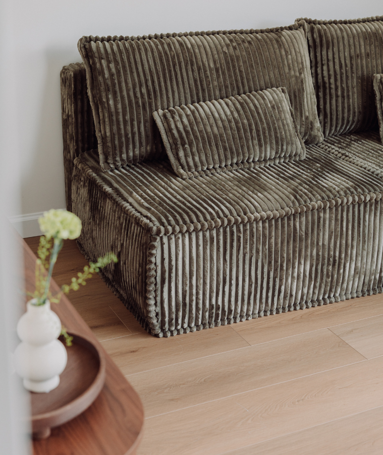 Tapicerowana sofa w prążkowanej, brązowej tkaninie z dekoracyjnymi poduszkami; ustawiona na jasnej drewnianej podłodze, obok niski stolik z wazonem i kwiatami w minimalistycznym wnętrzu.