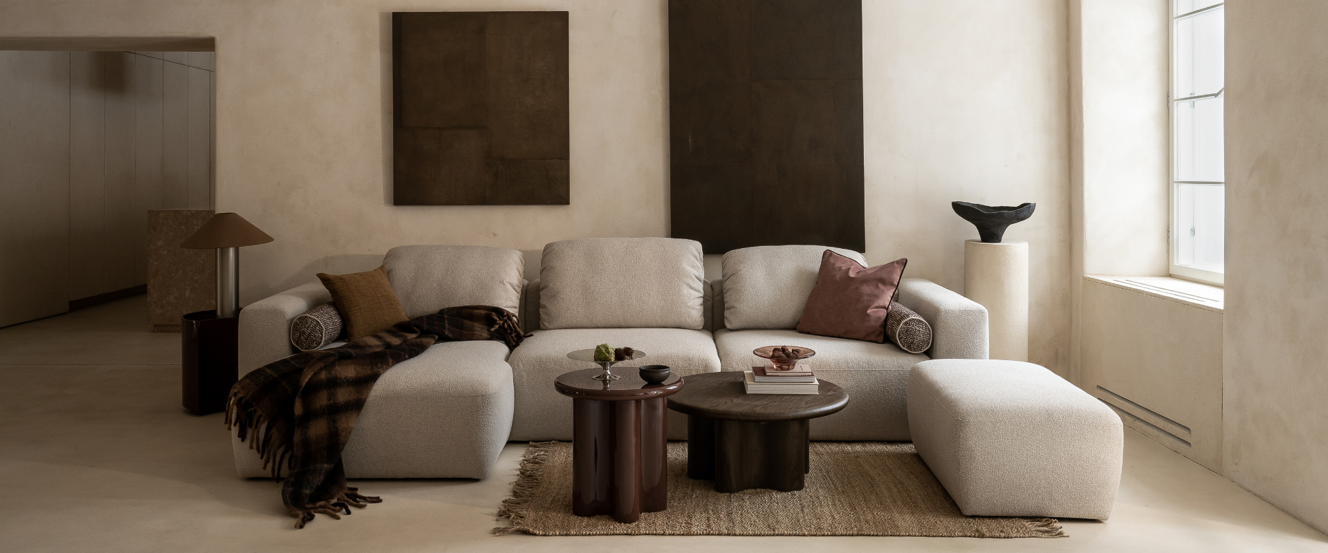 Przestronny salon w ciepłych, beżowo-brązowych tonach: modułowa sofa z szezlongiem i pufą, dwa niskie stoliki kawowe z ciemnego drewna, minimalistyczne obrazy na ścianie oraz duże okno wpuszczające miękkie, naturalne światło.