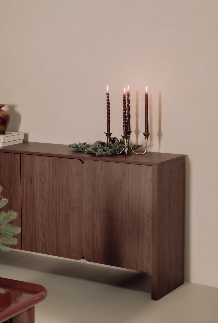 Drewniana komoda w minimalistycznym wnętrzu, na blacie metalowy świecznik z zapalonymi świecami i gałązkami świerku, tworzące nastrojową, świąteczną aranżację.