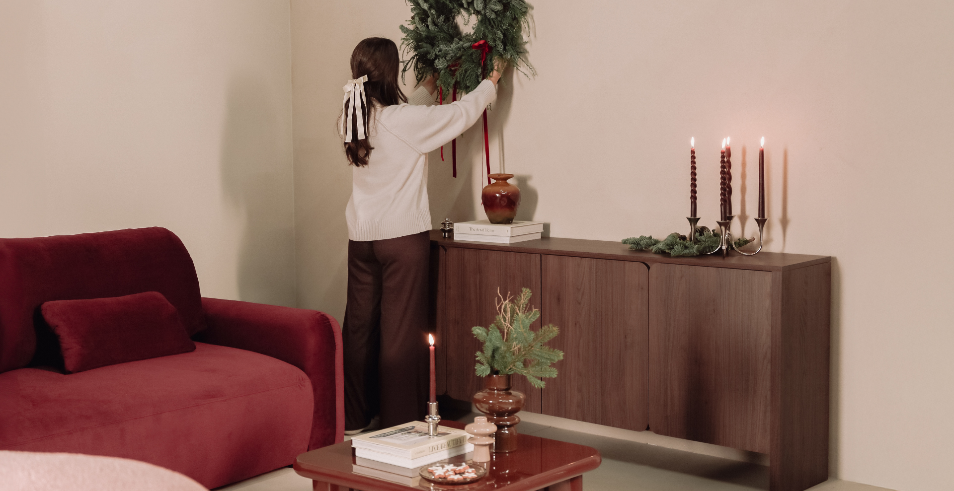 Kobieta dekoruje ścianę świątecznym wiankiem nad drewnianą komodą, na której stoją zapalone świece i dekoracje; w salonie widoczna czerwona sofa, stolik kawowy i bożonarodzeniowe ozdoby w ciepłej, nastrojowej aranżacji.