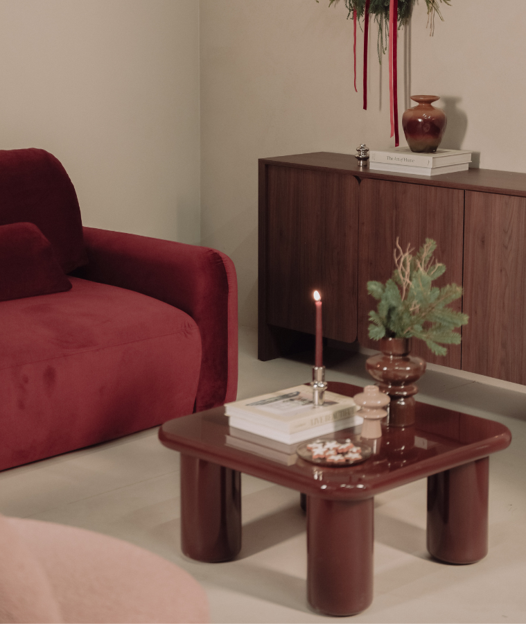 Przytulny salon w ciepłych barwach z czerwonym fotelem, drewnianą komodą i niskim stolikiem kawowym; na stoliku zapalona świeca, książki i świąteczne dekoracje, tworzące nastrojową aranżację.